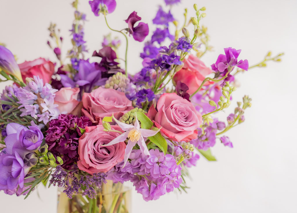 close-up of A flower arrangement in a glass container in front of a white backdrop on a small table. It's filled with lavender and purple flowers, including rose, stock, delphinium, and statice, hyacinth, and clematis.
