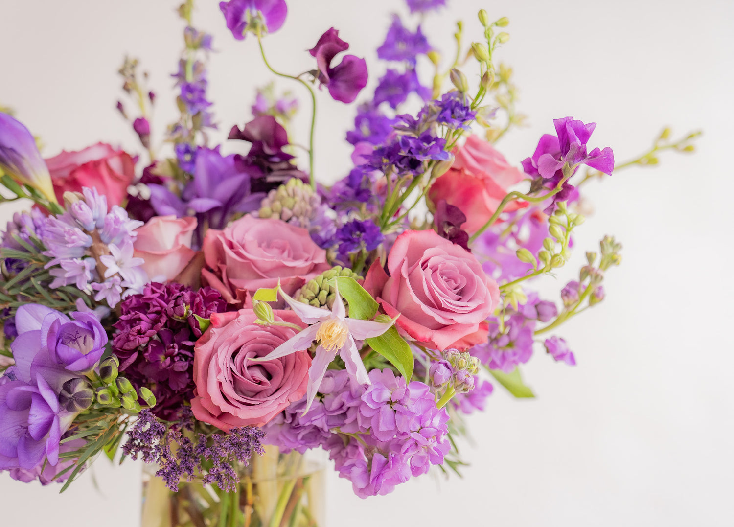 close-up of A flower arrangement in a glass container in front of a white backdrop on a small table. It's filled with lavender and purple flowers, including rose, stock, delphinium, and statice, hyacinth, and clematis.