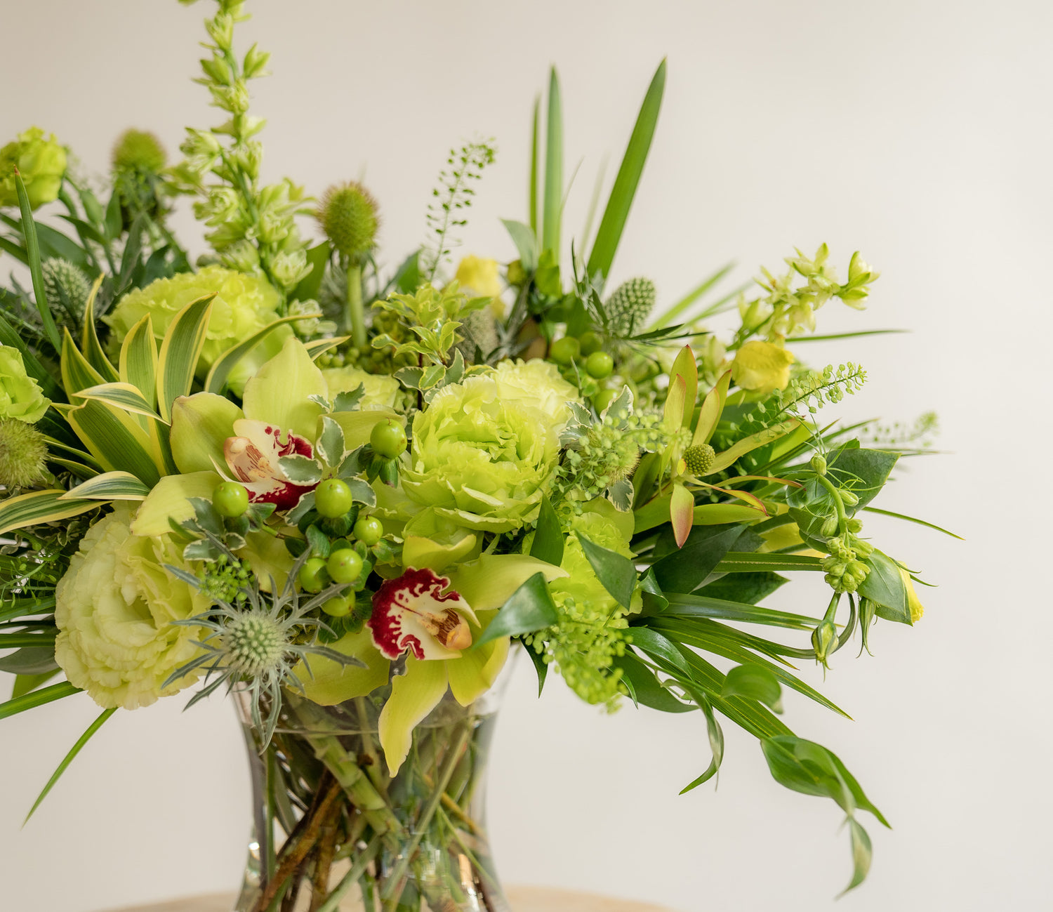 closeup of A flower arrangement in a glass container in front of a white backdrop on a small table. It's filled with green and chartreuse flowers and foliage, including rose, lisianthus, eucalyptus, and pennycrest, cymbidium orchid.