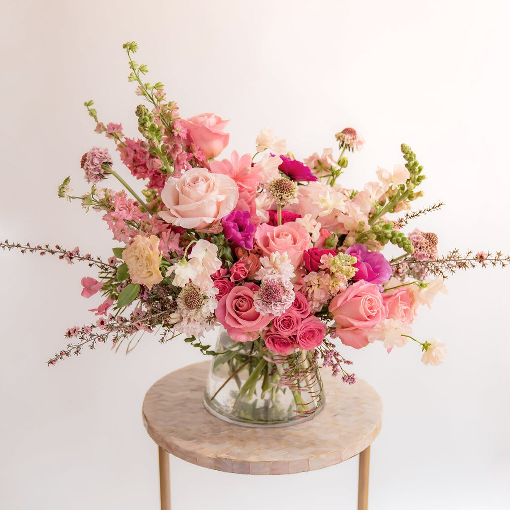 A flower arrangement in a glass container in front of a white backdrop on a small table. It's filled with pink and magenta flowers, including rose, scabiosa, sweet pea, snapdragon, and delphinium.