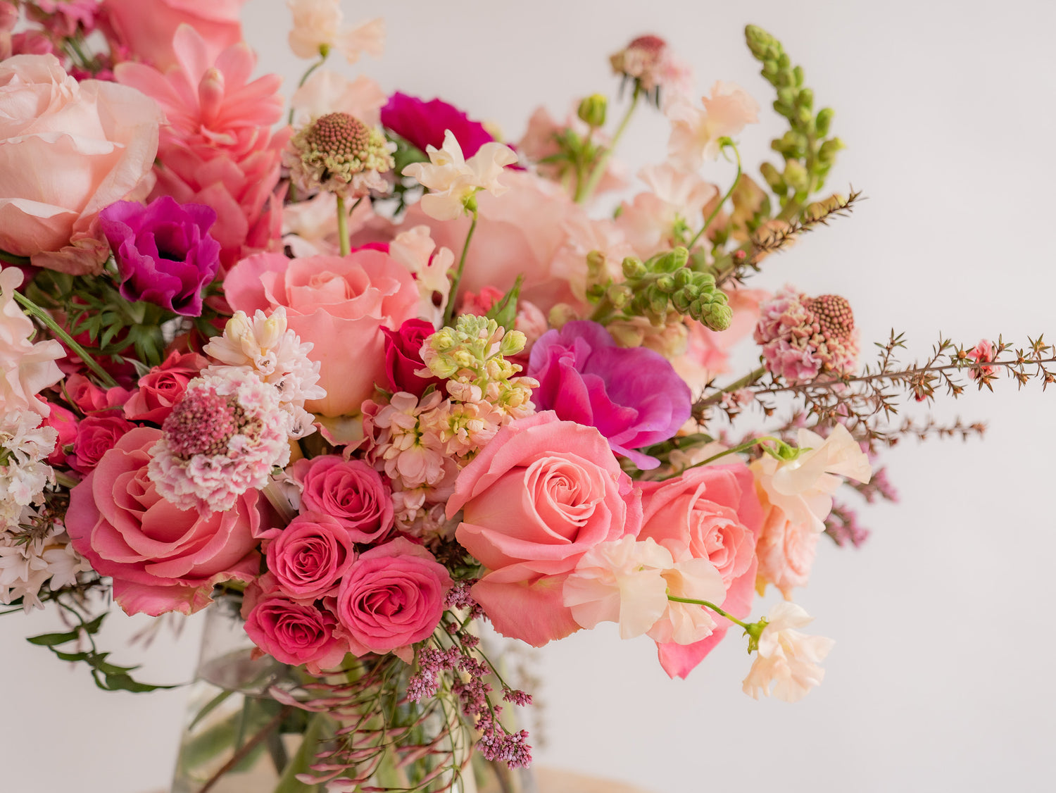 closeup of A flower arrangement in a glass container in front of a white backdrop on a small table. It's filled with pink and magenta flowers, including rose, scabiosa, sweet pea, snapdragon, and delphinium.