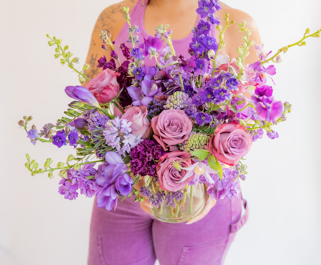a woman in all purple holds A flower arrangement in a glass container in front of a white backdrop. It's filled with lavender and purple flowers, including rose, stock, delphinium, and statice, hyacinth, and clematis.