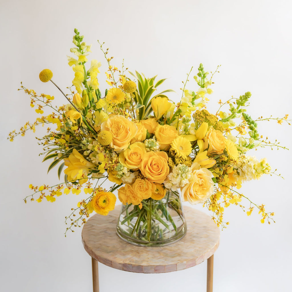 A flower arrangement in a glass container in front of a white backdrop on a small table. It's filled with yellow and gold flowers, including rose, snapdragon, billy balls, spray rose, yarrow, stock, daffodils, and oncidium orchid.