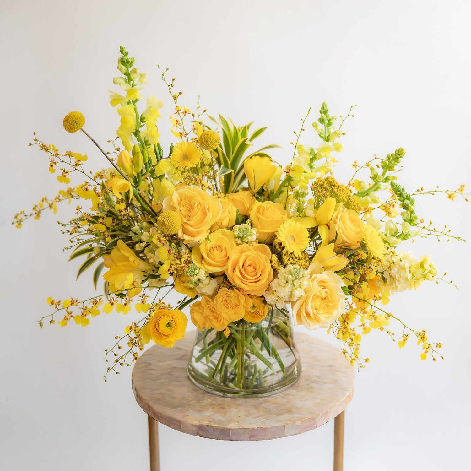 A flower arrangement in a glass container in front of a white backdrop on a small table. It's filled with yellow and gold flowers, including rose, snapdragon, billy balls, spray rose, yarrow, stock, daffodils, and oncidium orchid.
