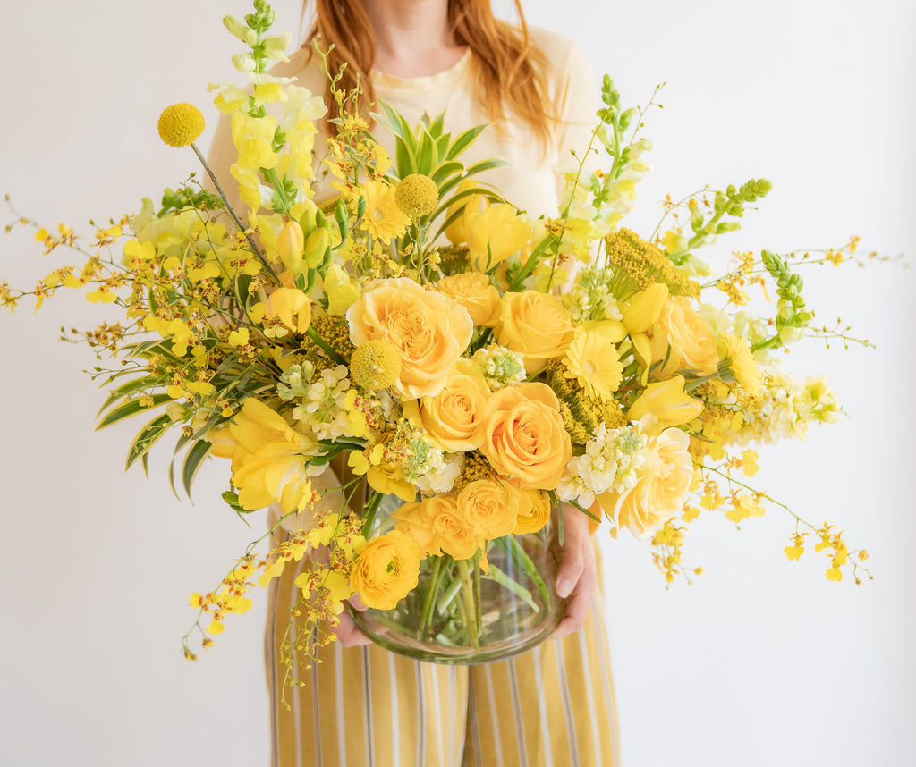 A woman holding a monochromatic flower arrangement in a glass container in front of a white backdrop. It's filled with yellow and gold flowers, including rose, snapdragon, billy balls, spray rose, yarrow, stock, daffodils, and oncidium orchid.