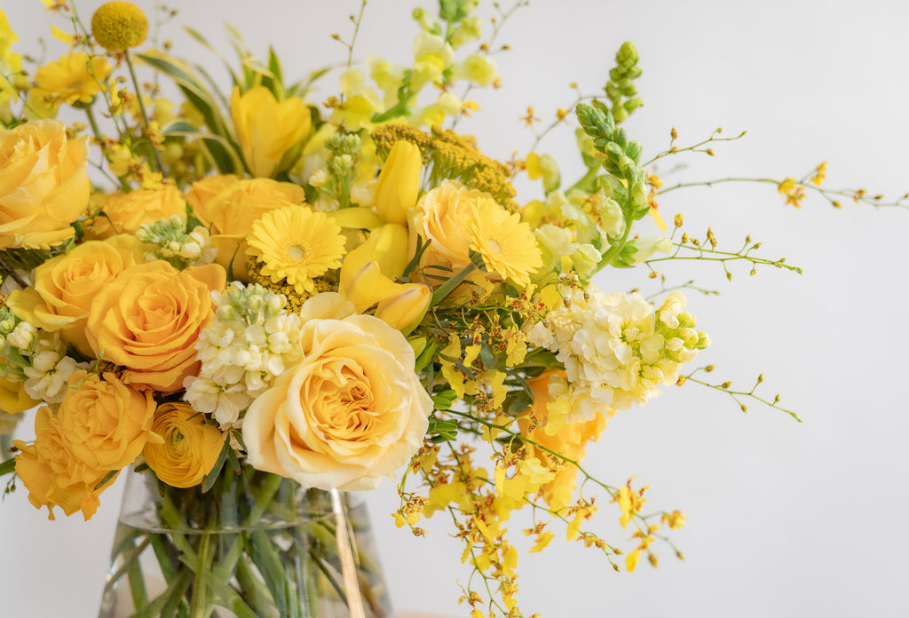 A flower arrangement in a glass container in front of a white backdrop on a small table. It's filled with yellow and gold flowers, including rose, snapdragon, billy balls, spray rose, yarrow, stock, daffodils, and oncidium orchid.