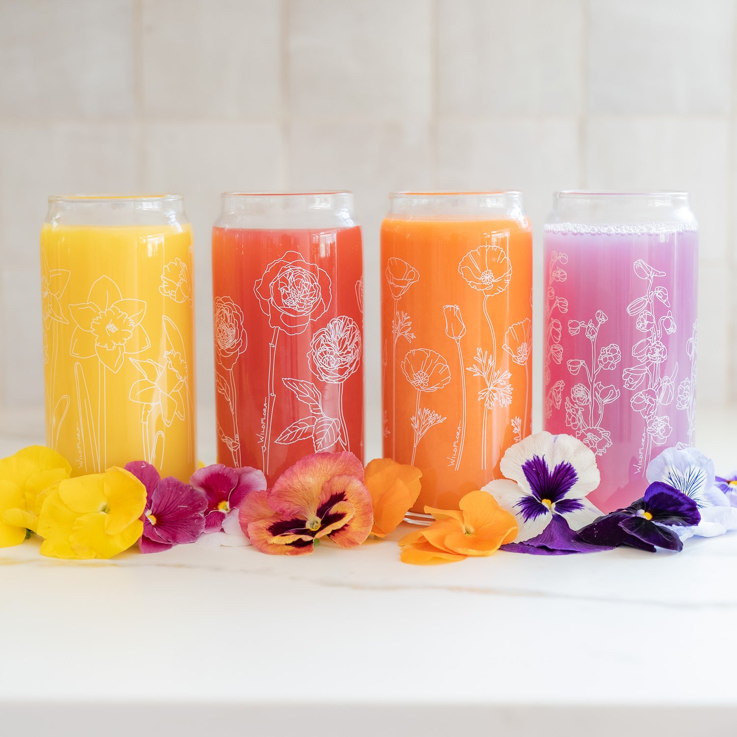 A set of 4 botanically-designed, beer-can shaped tumblers on a counter, adorned with pansy blooms and filled with coral, orange, yellow, and purple beverages. One tumbler has roses printed on it, another poppies, another daffodils, and the last delphinium.