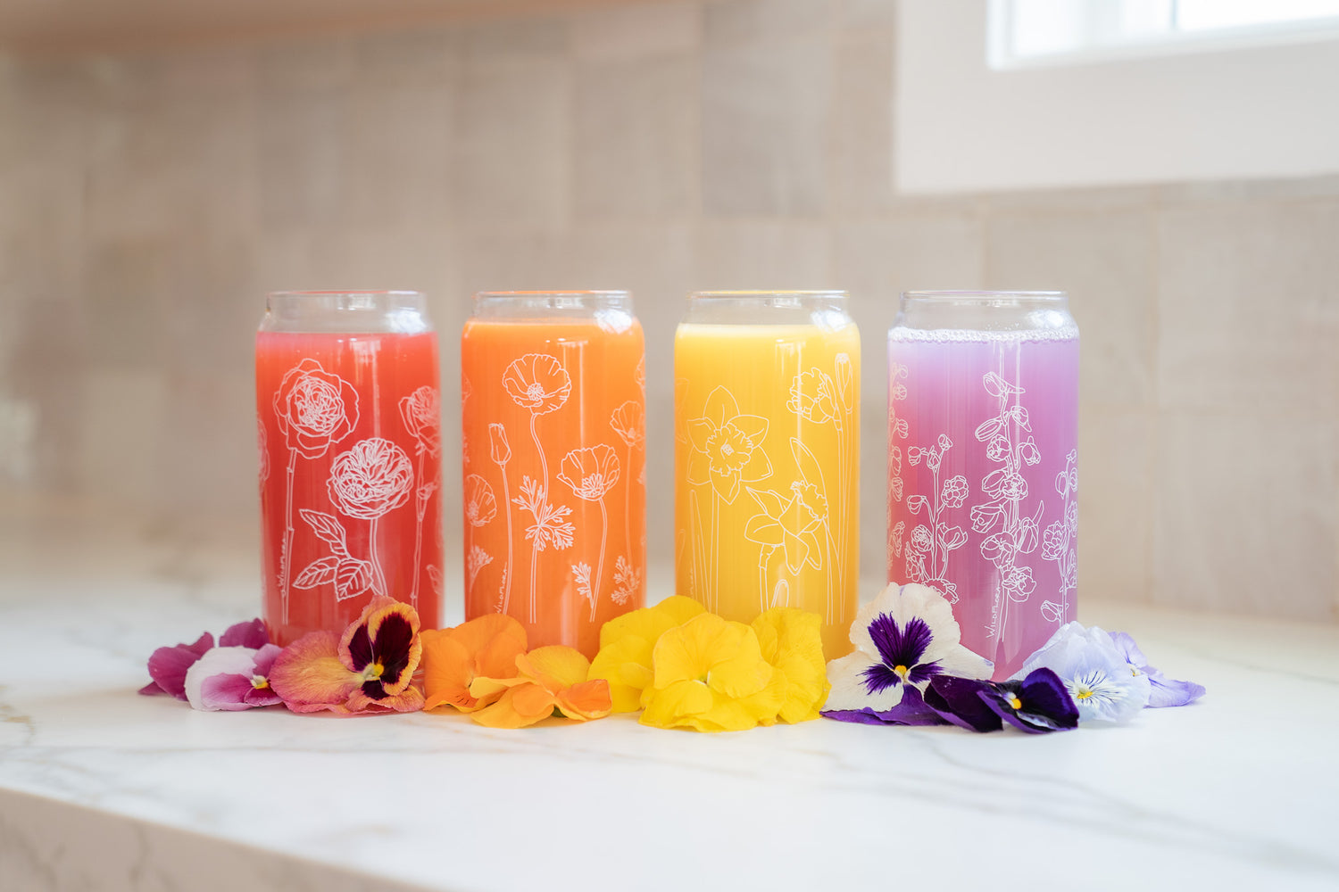 A set of 4 botanically-designed, beer-can shaped tumblers on a counter, adorned with pansy blooms and filled with coral, orange, yellow, and purple beverages. One tumbler has roses printed on it, another poppies, another daffodils, and the last delphinium.