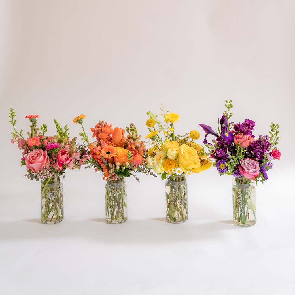 A set of 4 botanically-designed, beer-can shaped tumblers in front of a white backdrop. One glass has pink flowers, the next orange, the next yellow, and the last purple, including rose, piccolini daisy, iris, billy ball, chamomile, tulip, snapdragon, and straw flowers blooms
