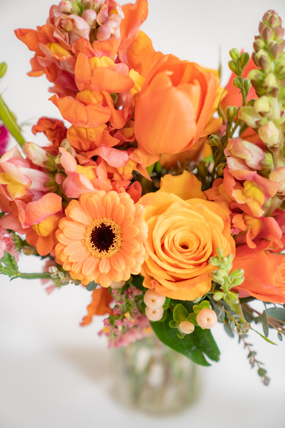 closeup of orange flowers, including rose, tulip, snapdragon, hypericum berries, and piccolini daisy.