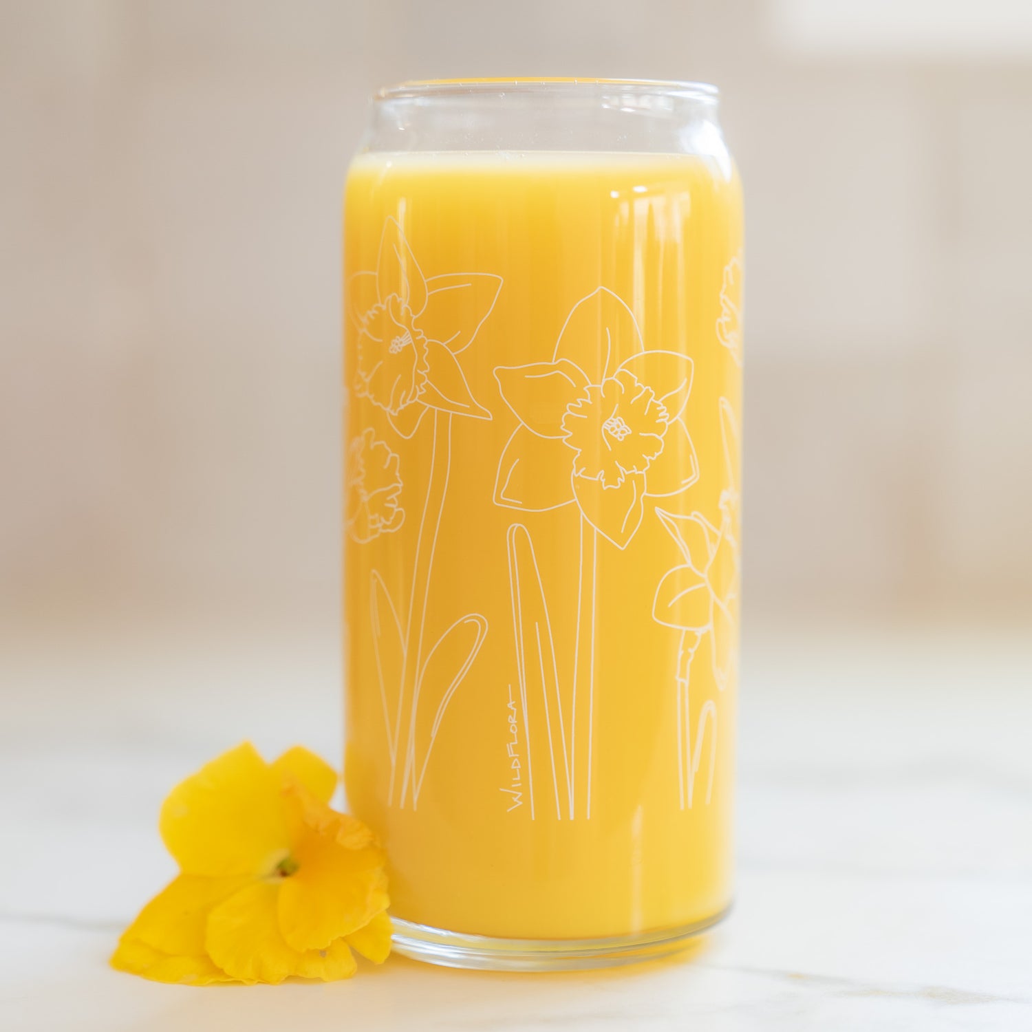 A clear 16 ounce tumbler with a white daffodil graphic design. The tumbler is beer-can shaped, adorned by a yellow pansy bloom, and filled with a yellow drink.