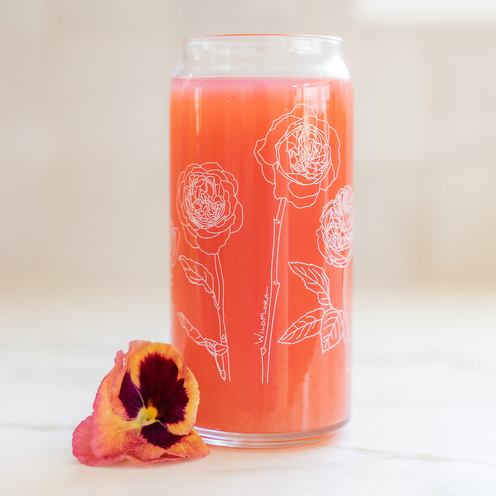 A clear 16 ounce tumbler with a white garden rose graphic design. The tumbler is beer-can shaped, adorned by a pink and purple pansy bloom, and filled with a coral-colored drink.