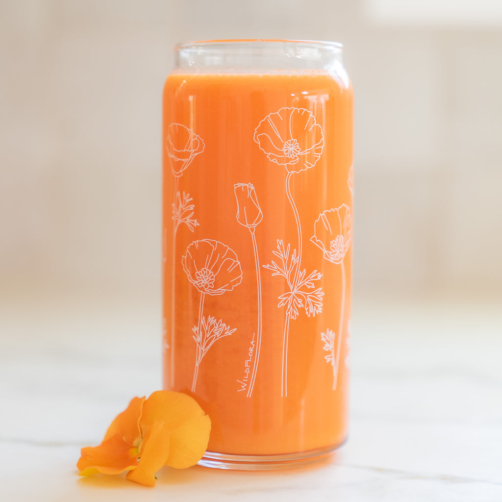 A clear 16 ounce tumbler with a white california poppy graphic design. The tumbler is beer-can shaped, adorned by a gold pansy bloom, and filled with an orange drink.