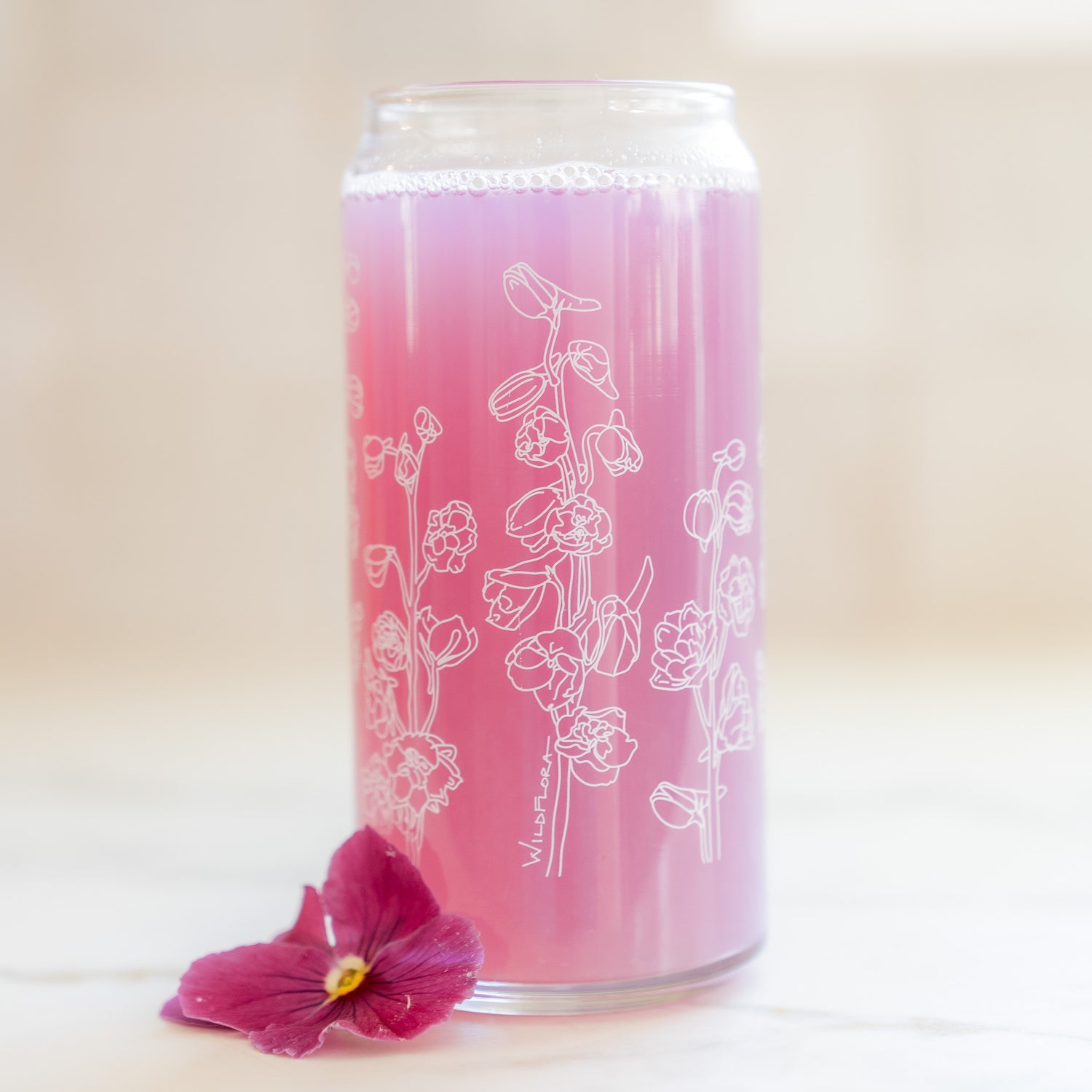 A clear 16 ounce tumbler with a white delphinium graphic design. The tumbler is beer-can shaped, adorned by a fuchsia pansy bloom, and filled with a purple drink.