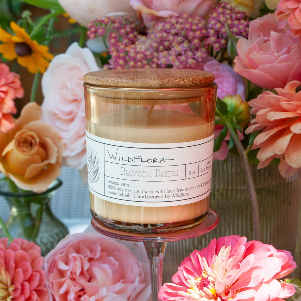 Blossom Breeze single-wick, white soy wax candle in a tan glass container with a wood lid. The label is apothecary style and shows the scent. Behind the candle are pink flowers.