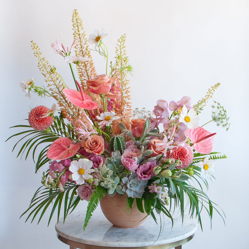 A very large pink, peach, white, and green flower arrangement sits on a marble table. The design is inspired by both the desert and lush vibes of Palm Springs, California, and includes cacti, succulents, roses, anthurium, cosmos, lisianthus, Queen Anne's lace, foxtail lily, orchids, snapdragon, palm fronds, and monstera leaves in a round terracotta pot.