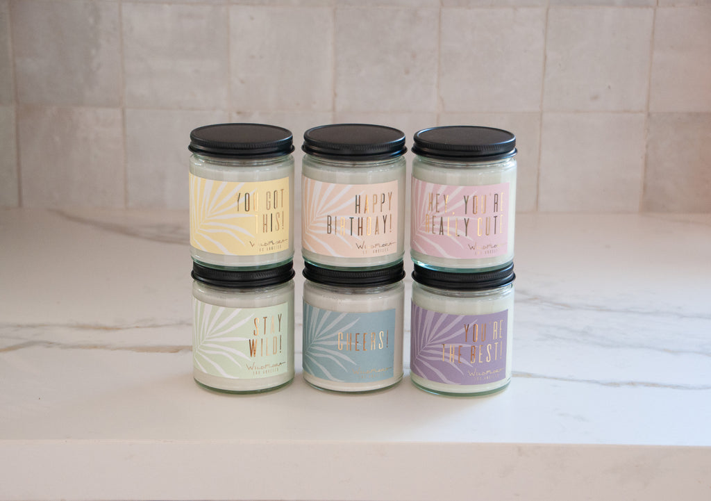 6 Expression candles sitting on a marble counter. They say "you're the best!" on a yellow label, "happy birthday!" on a peach label, "hey, you're really cute," on a pink label, "stay wild!" on a light green label, "cheers!" on a light blue label, and "you're the best" on a light purple label.