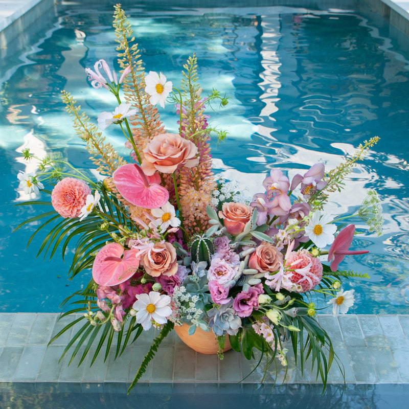 A very large pink, peach, white, and green flower arrangement sits near a pool. The design is inspired by both the desert and lush vibes of Palm Springs, California, and includes cacti, succulents, roses, anthurium, cosmos, lisianthus, Queen Anne's lace, foxtail lily, orchids, snapdragon, palm fronds, and monstera leaves in a round terracotta pot.