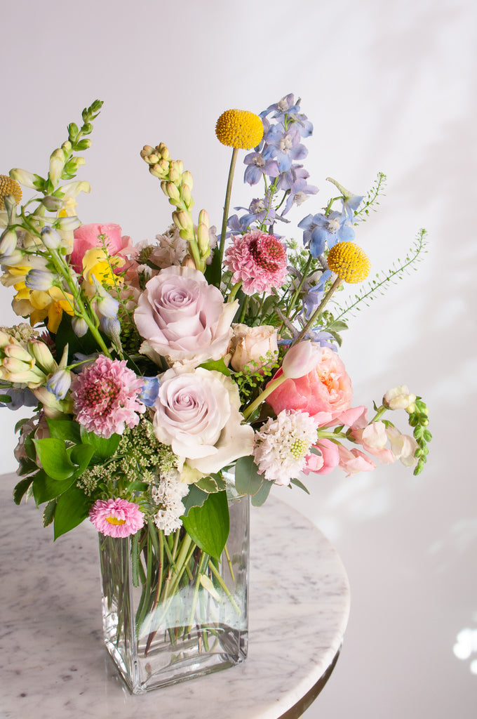 A glass vase with a small flower arrangement sits on a marble table in front of a white backdrop.  The flowers are pastel purple, pink, peach, coral, and yellow, and blue with greens spilling around. The flowers include billy ball, rose, scabiosa, hyacinth, delphinium, tulip, snapdragon, and aster.