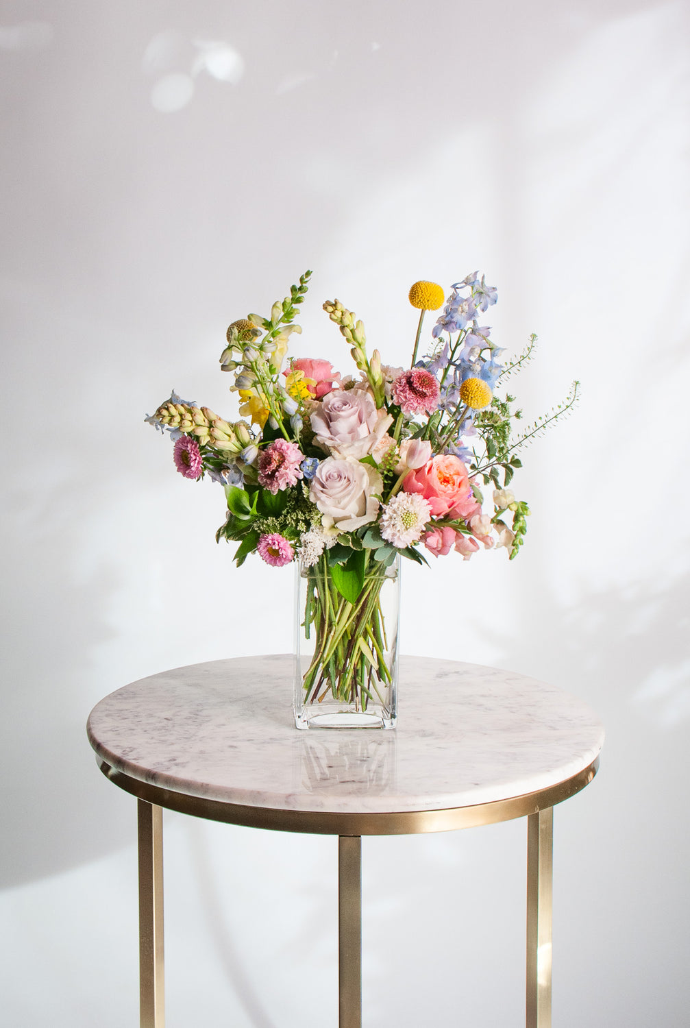 A glass vase with a small flower arrangement sits on a marble table in front of a white backdrop.  The flowers are pastel purple, pink, peach, coral, and yellow, and blue with greens spilling around. The flowers include billy ball, rose, scabiosa, hyacinth, delphinium, tulip, snapdragon, and aster.