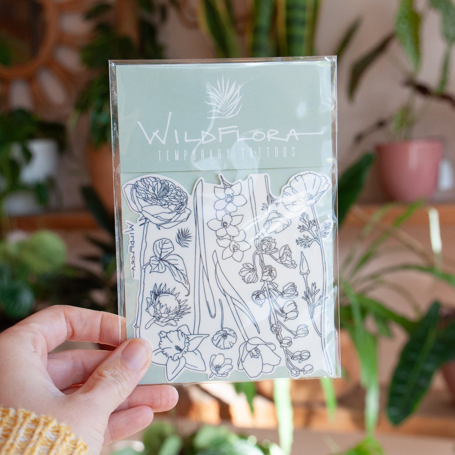 Hand holding a package of temporary tattoos that are black line drawings of varying flowers, including rose, protea, daffodil, California poppy, tulip, orchid, and delphinium.
