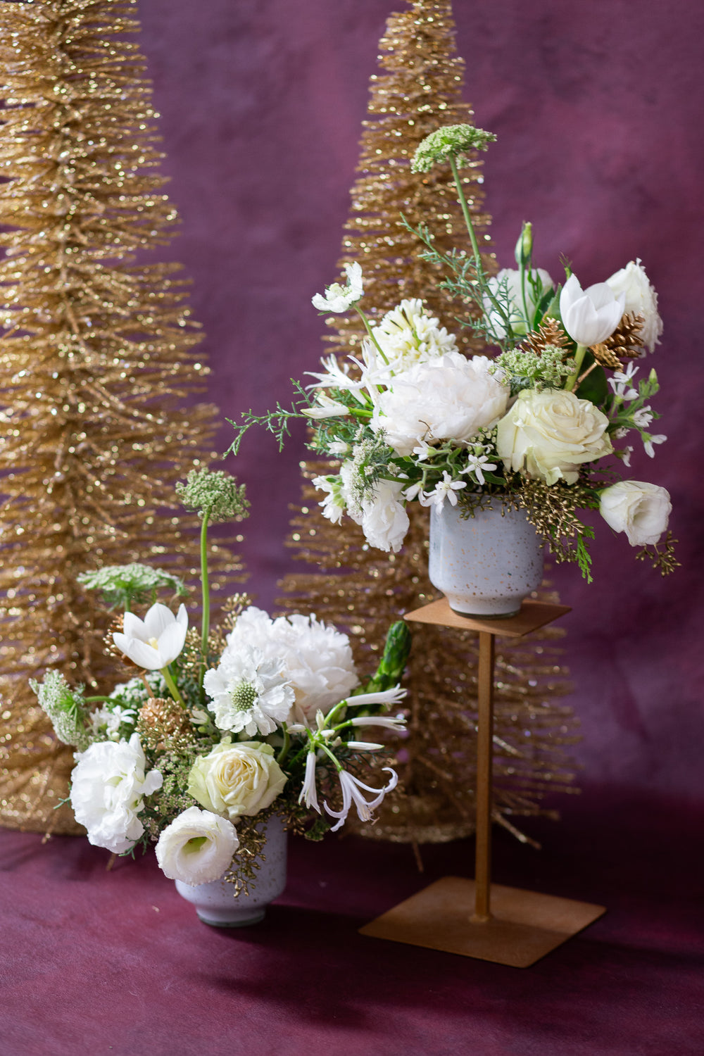 WildFlora's Frost-Kissed Arrangement Pair on in front of a maroon backdrop, accompanied by gold Christmas trees. They have green, white, and gold flowers, and include rose, Queen Anne's lace, pine cones, lily, scabiosa, tulip, and peony.