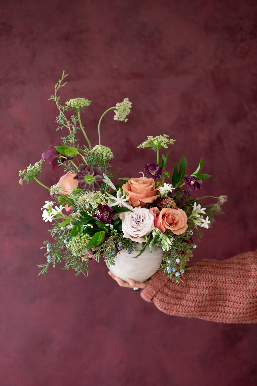 WildFlora's Winter's Garden Arrangement in a spherical grey vase, held by a hand in front of a maroon backdrop. It has sienna orange, pastel violet, red, pink, fuchsia, eggplant, and white flowers, and includes rose, scabiosa, hellebore, Queen Anne's lace, and juniper.