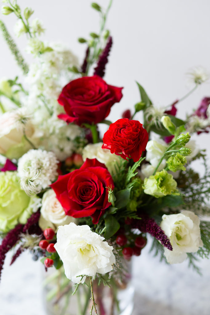 WildFlora's Holiday Cheer Arrangement in a glass vase on a small marble table in front of a white backdrop. It has red, green, white, and chartreuse flowers, and includes rose, delphinium, snapdragon, scabiosa, piccolini daisy, berries, and lisianthus.