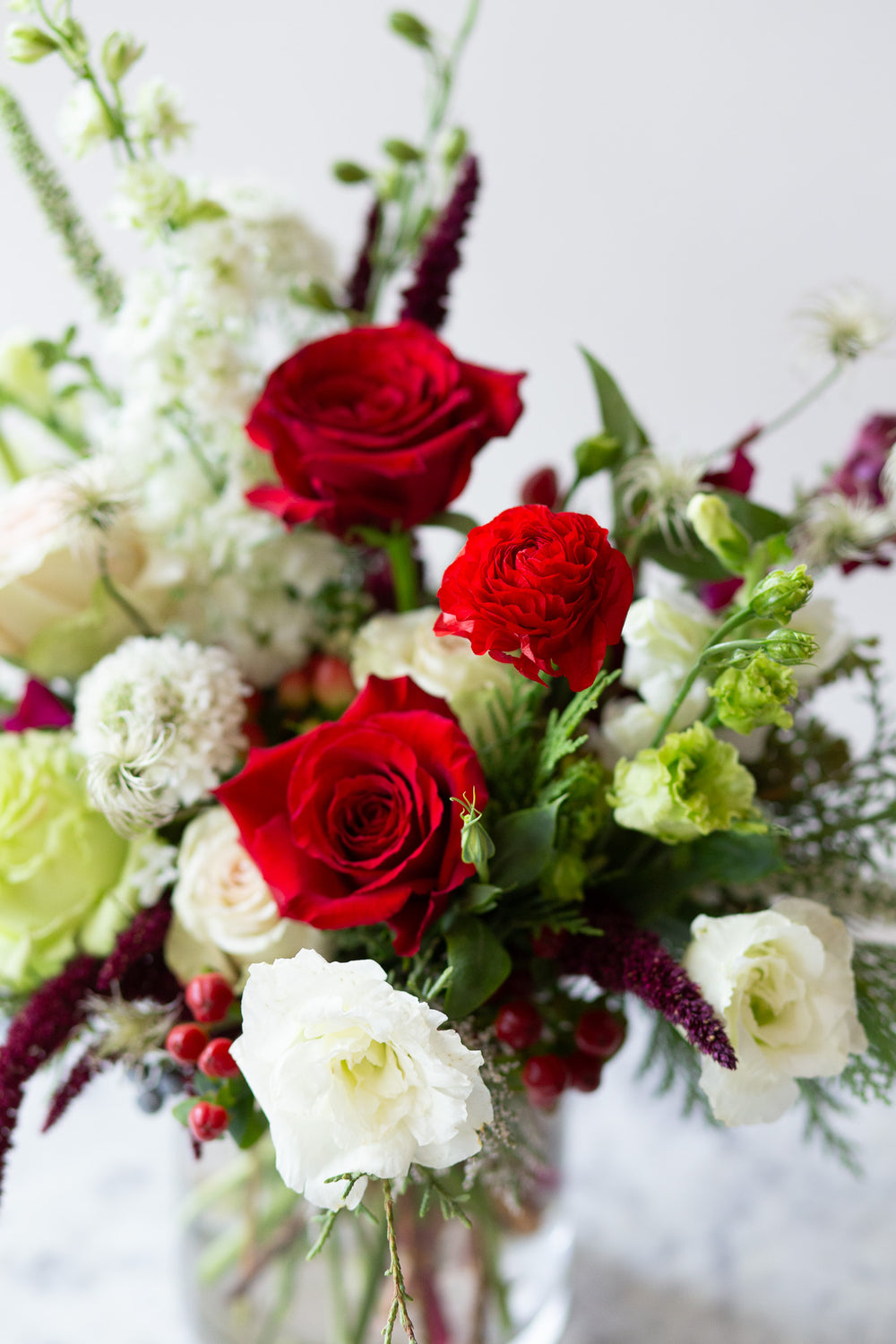 WildFlora's Holiday Cheer Arrangement in a glass vase on a small marble table in front of a white backdrop. It has red, green, white, and chartreuse flowers, and includes rose, delphinium, snapdragon, scabiosa, piccolini daisy, berries, and lisianthus.