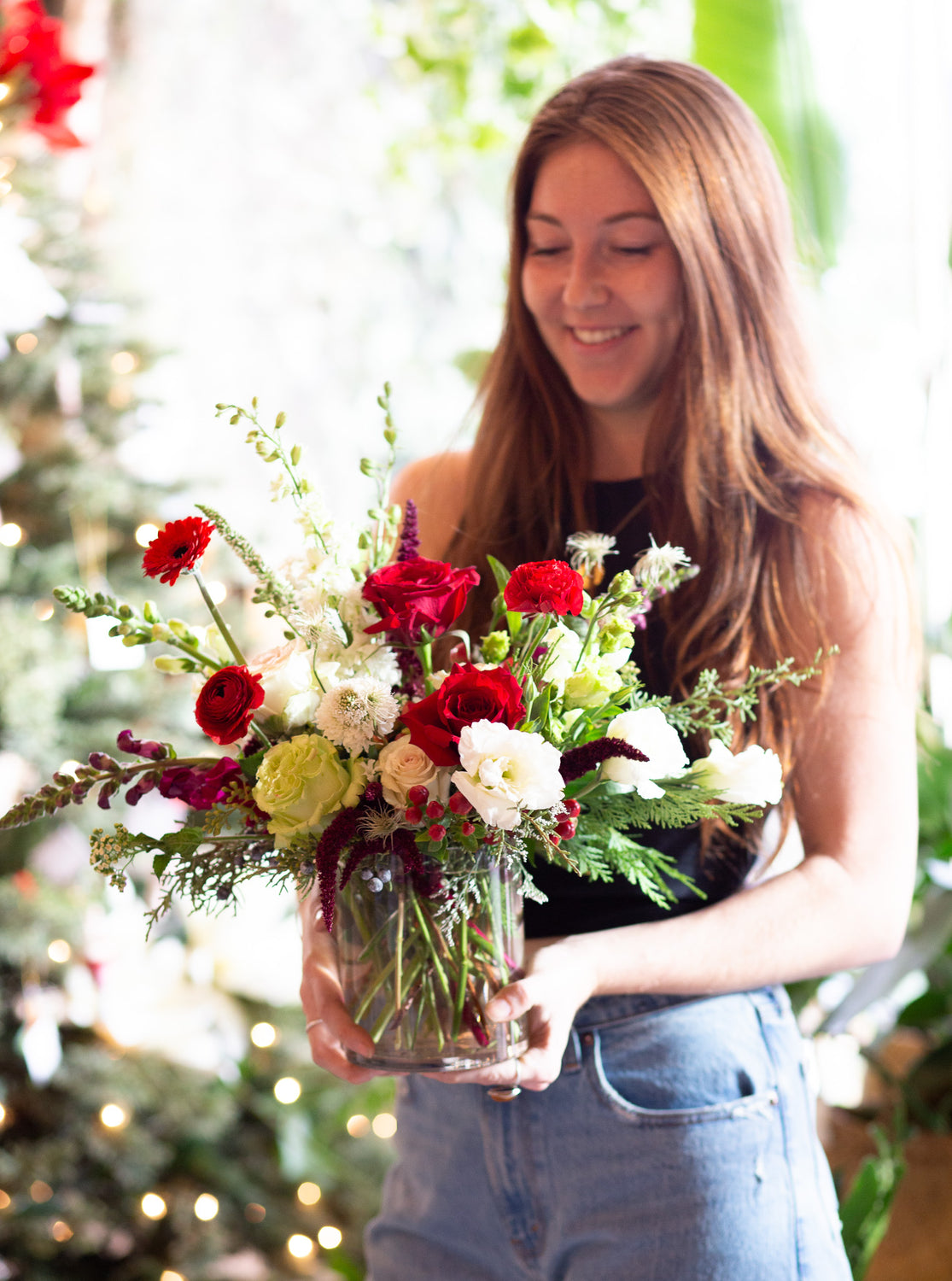 A woman holds WildFlora's Holiday Cheer Arrangement in a glass vase. It has red, green, white, and chartreuse flowers, and includes rose, delphinium, snapdragon, scabiosa, piccolini daisy, berries, and lisianthus.