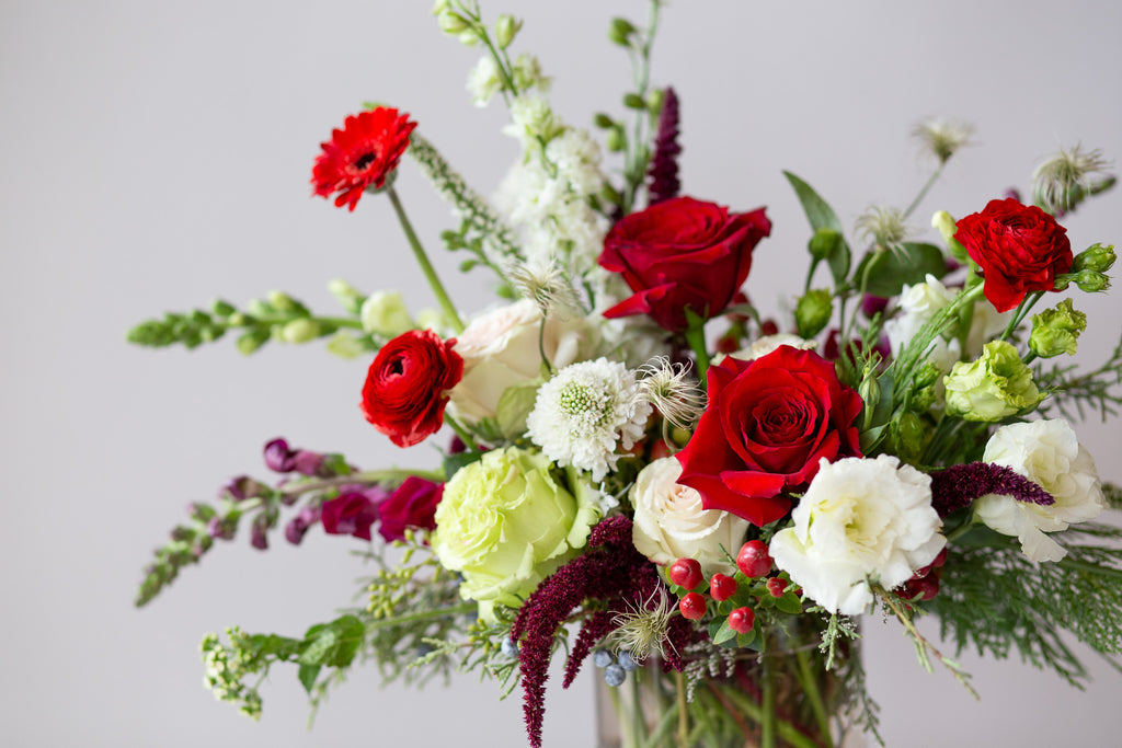 WildFlora's Holiday Cheer Arrangement in a glass vase on a small marble table in front of a white backdrop. It has red, green, white, and chartreuse flowers, and includes rose, delphinium, snapdragon, scabiosa, piccolini daisy, berries, and lisianthus.