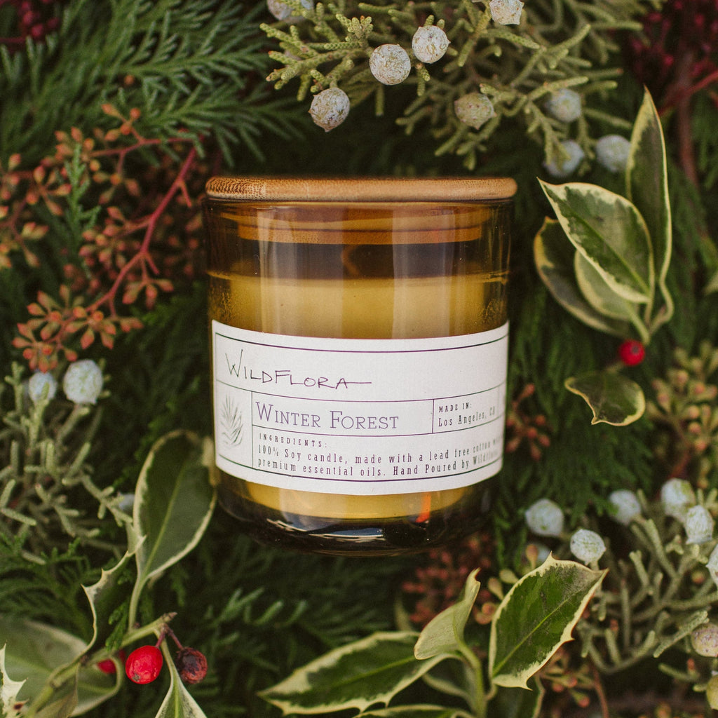WildFlora Winter Forest Candle jar with a label and wood lid on a bed of winter greens