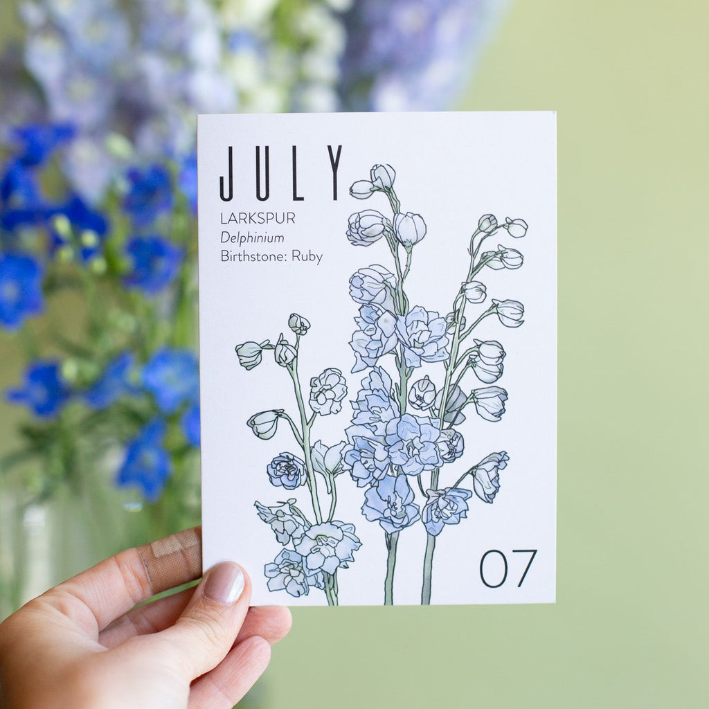 A hand holds a July birth month birthday card that has artwork of delphinium on it and also lists the birth month gemstone. blurry in the background are blue delphinium blooms