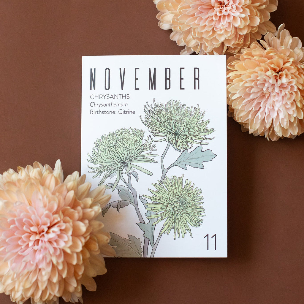 A November birth month card that has artwork of chrysanthemums on it and also lists the birth month gemstone. It lays on a brown background with three peach chrysanthemums around it.