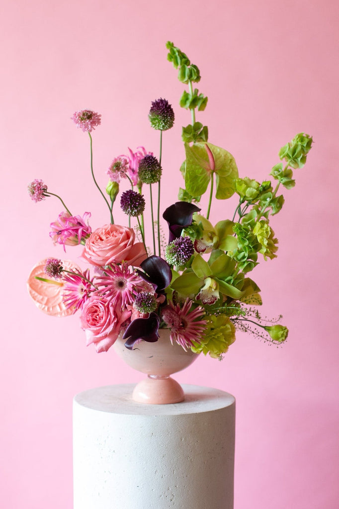 Pink & Green floral arrangement in a pink vase on a white pedestal against a pink background - very Glinda-Elphaba vibes