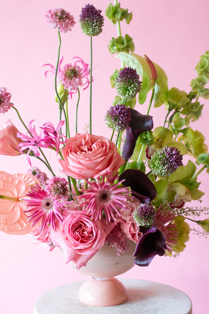 Pink & Green floral arrangement in a pink vase on a white pedestal against a pink background - very Glinda-Elphaba vibes