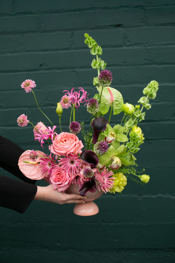 hands holding a Pink & Green floral arrangement in a pink vase against a dark green brick wall - very Glinda-Elphaba vibes