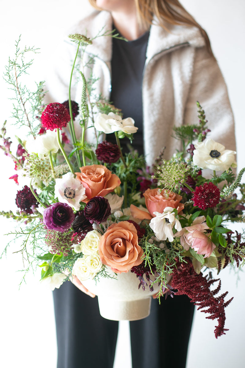woman holding A Floral arrangement with salmon-colored Moab Roses, white Queen Anne's Lace & anemone & Veronica, dark purple ranunculus & scabiosa, maroon amaranthus, pink hellebore, violet snapdragon with evergreen foliage in a white ceramic compote vase in front of a white green backdrop