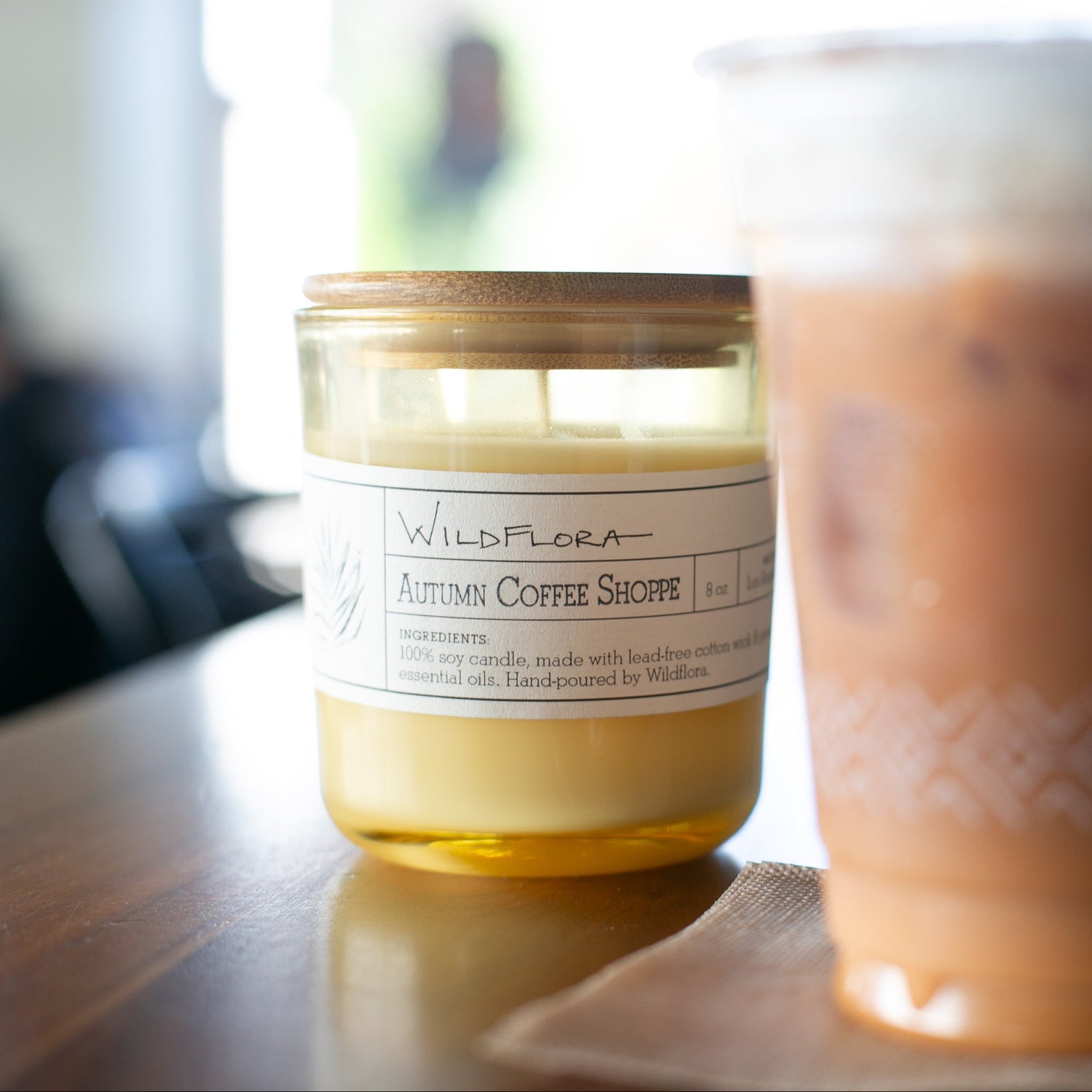 A jar candle labeled Wildflora 'Autumn Coffee Shoppe' sits on a wooden table next to an iced coffee cup in a softly blurred cafe setting.