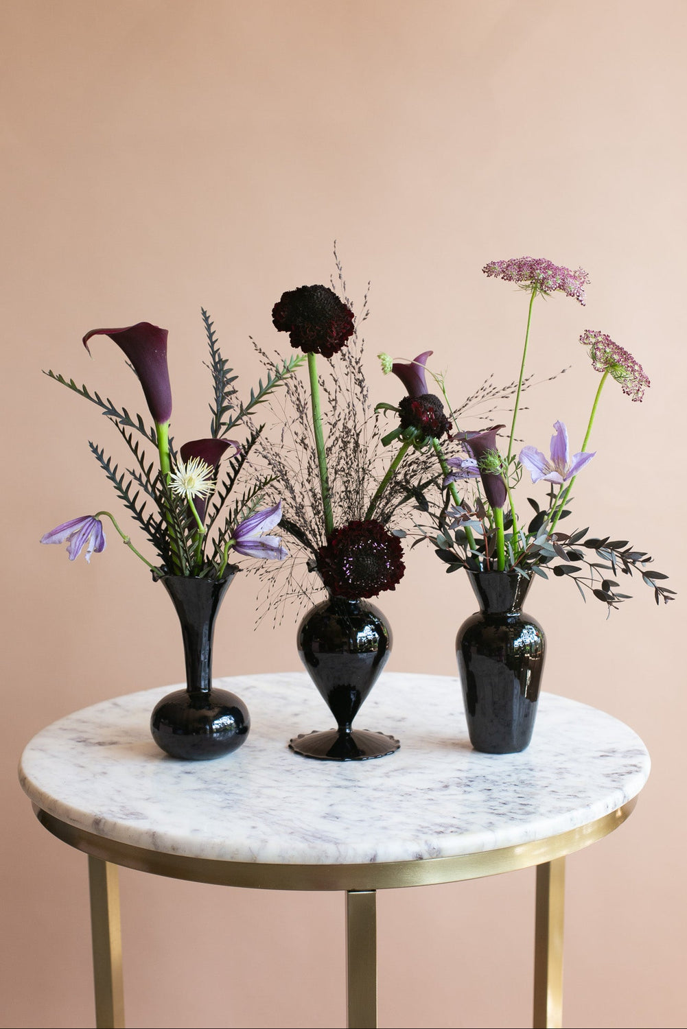 Three black glass vases filled with dark purple and lavender floral arrangements, displayed on a round white marble-top side table with gold legs against a warm beige wall.