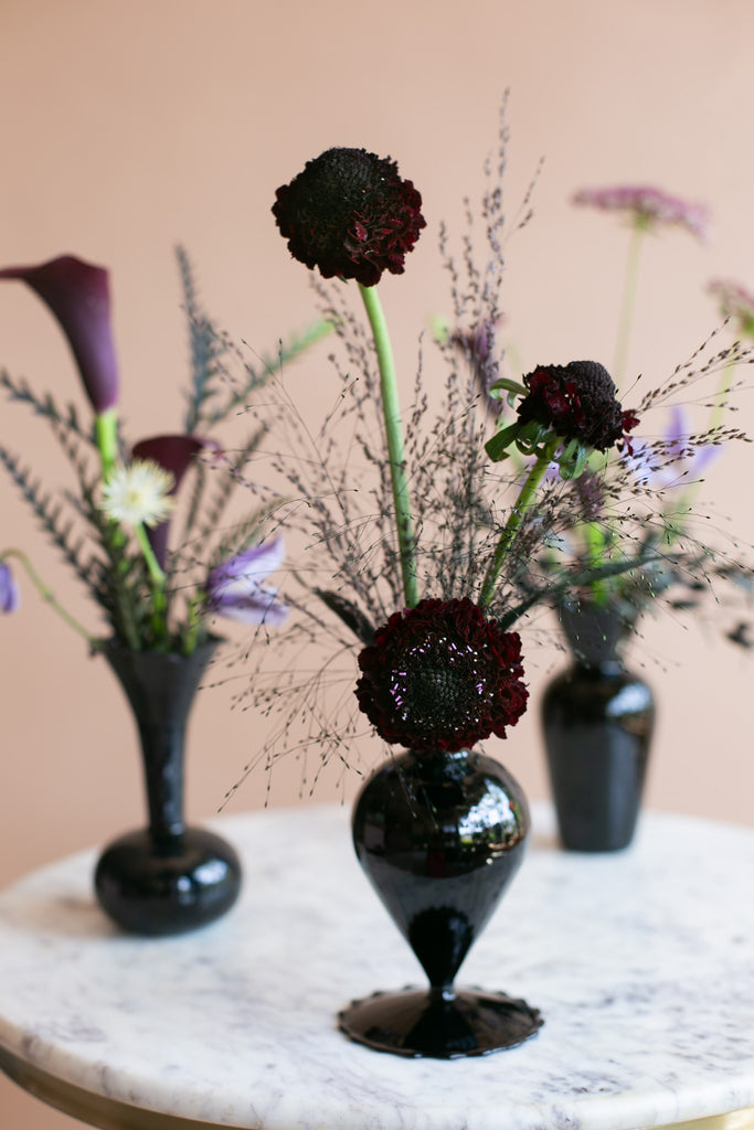 Three black glass vases with floral arrangements on a white marble table against a warm beige background. The center vase features dark burgundy flowers with feathery dried stems, flanked by purple calla lilies and lavender in the background vases.