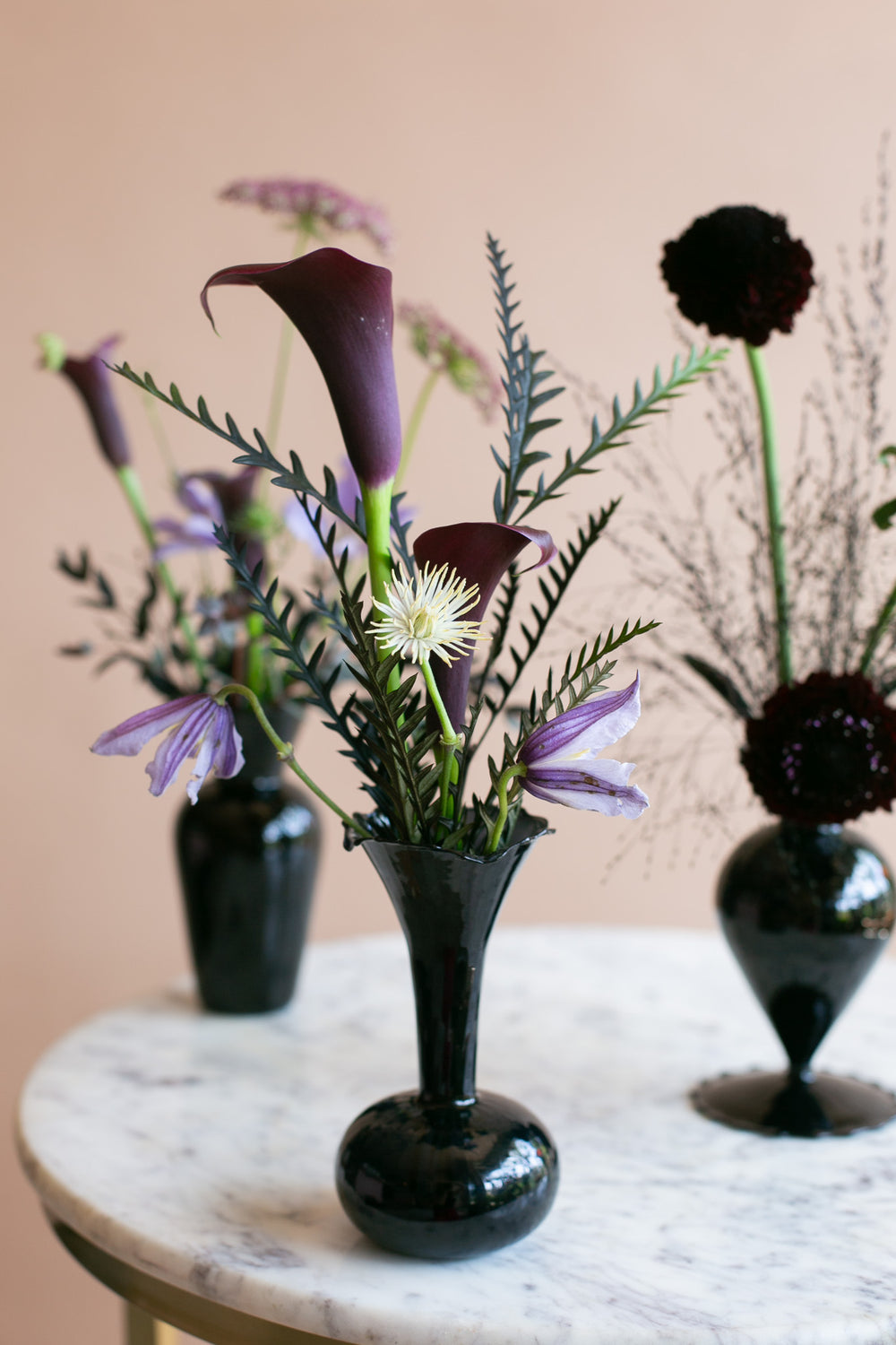 A dark glossy ceramic vase holding a moody floral arrangement with purple calla lilies, pale purple clements, dark burgundy blooms, and green foliage, displayed on a white marble-topped table against a warm beige background.