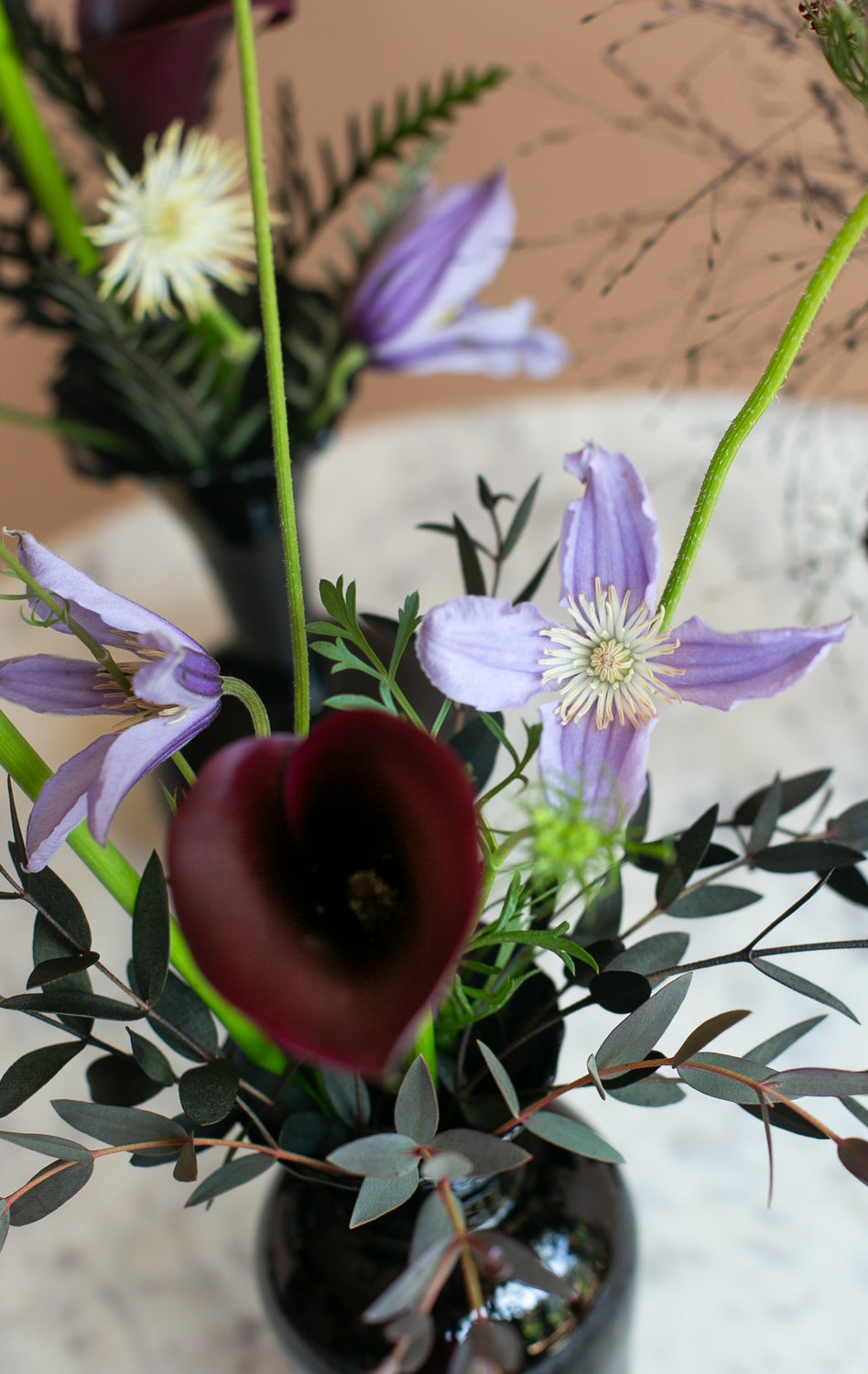 A close-up floral arrangement featuring light purple clematis flowers, a dark burgundy calla lily, and green foliage in a glossy black vase.