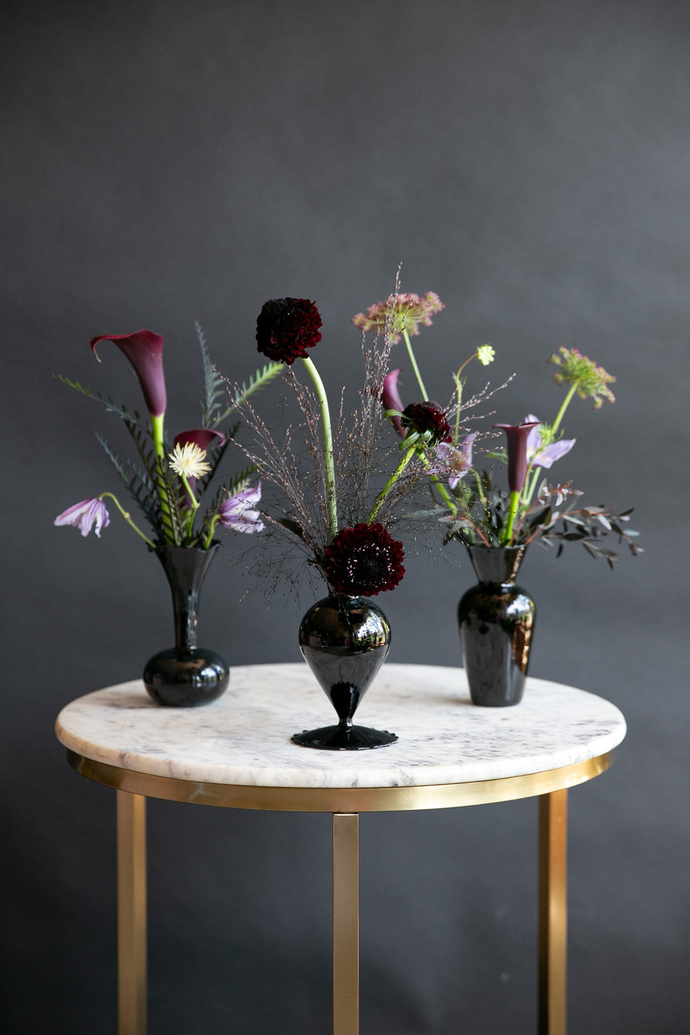 Three black glass vases containing dark, moody floral arrangements with burgundy calla lilies, deep red flowers, and green foliage, displayed on a round white marble and gold side table against a dark gray background.