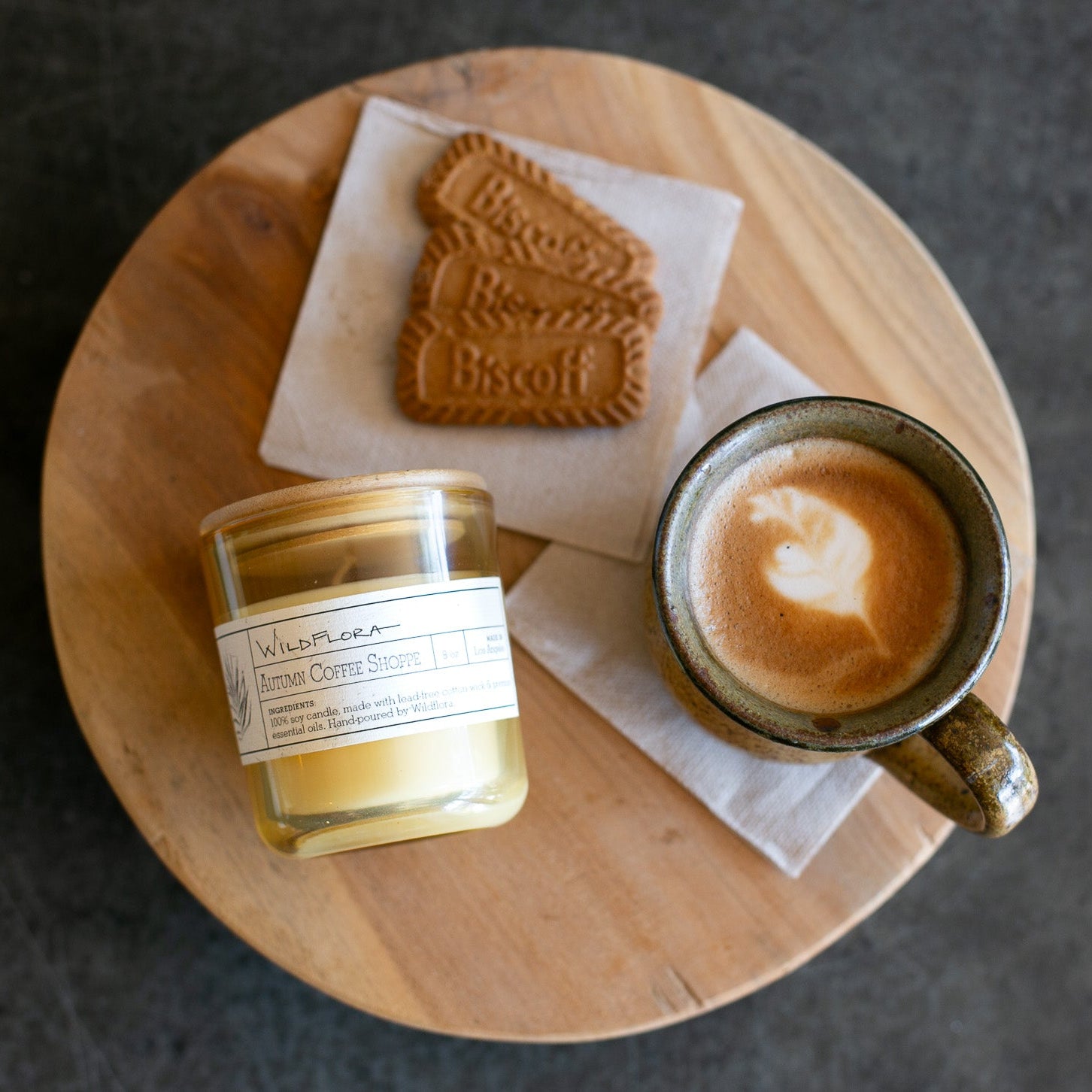 A styled overhead shot of a round wooden tray holding a candle in a glass jar, a ceramic mug of latte with latte art, and Biscoff cookies on white napkins, set against a dark gray background.