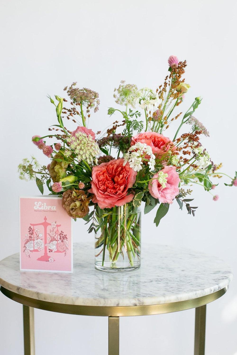 A vibrant wildflower arrangement in a clear glass vase, displayed on a white marble and gold side table next to a pink 'Libra' zodiac card against a white wall.