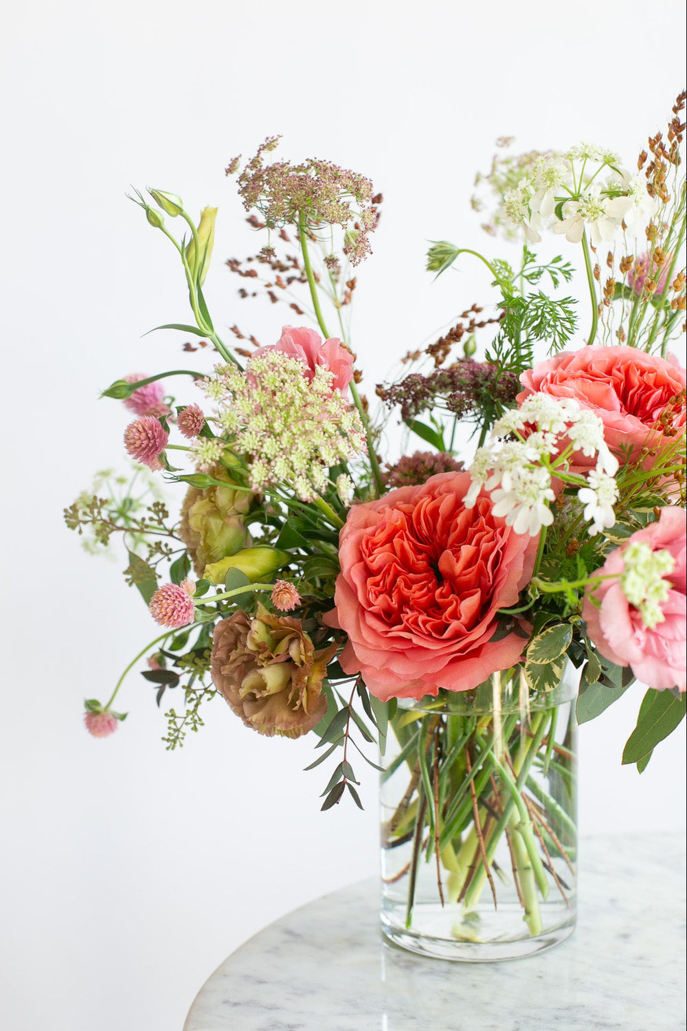 A vibrant floral arrangement featuring coral pink roses, Queen Anne's lace, and lisianthus in a clear glass vase, set on a white marble table against a clean white background.