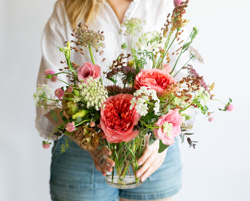 A person in a white button-up shirt and denim shorts holds a vibrant, wild flower arrangement in a clear glass vase against a plain white background.