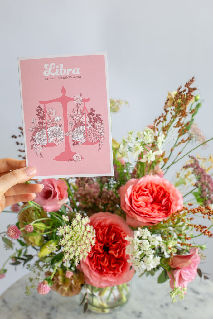 A hand holds a pink Libra astrology card in front of a vibrant floral arrangement with pink and coral roses, Queen Anne's lace, and green foliage against a white background.