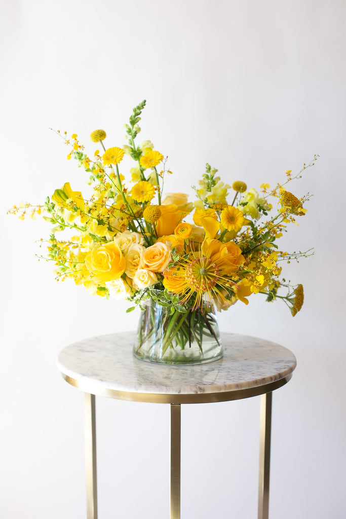 A flower arrangement in a glass container in front of a white backdrop on a small marble table. It's filled with yellow and gold flowers, including rose, billy balls, spray rose, yarrow, snap dragon, calla lily, and oncidium orchid. 
